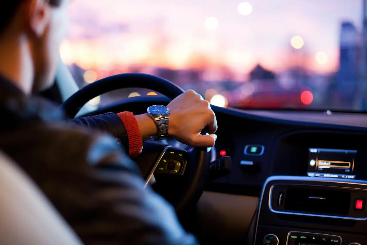 Motorista com a mão no volante dirigindo pela cidade ao entardecer